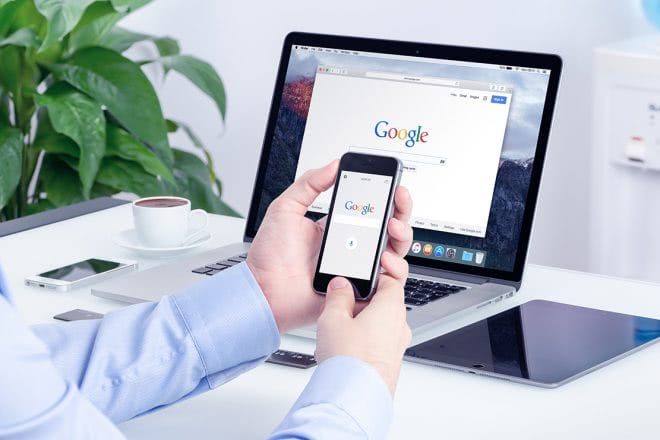 Hand mit Smartphone und Google-Suche, daneben ein Laptop